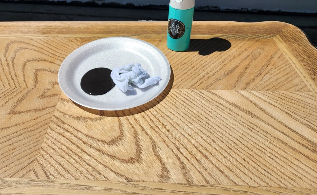 a paper plate with black paint and a rag, on a table where there is stain still present